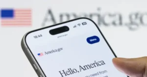 The image shows a smartphone displaying the America.gov website with a greeting that reads "Hello, America," set against a backdrop featuring the American flag.