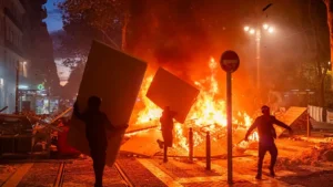 The image depicts a nighttime scene of a protest or riot, with individuals carrying large objects and a large fire burning in the background.