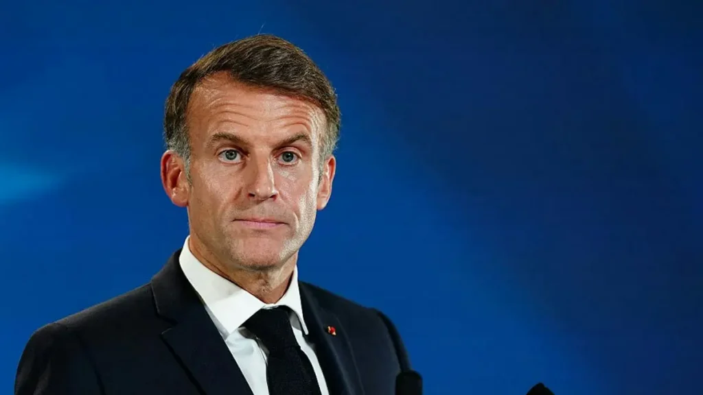 The image shows a man in a dark suit and tie, standing against a blue background, likely during a formal or official event.