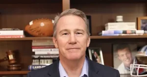 The image shows a man in a suit standing in front of a bookshelf filled with books, a football, a gavel, and a framed photo.