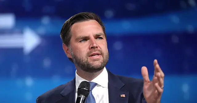 The image shows a man in a suit speaking into a microphone, gesturing with his hand, against a blue background with a white arrow.