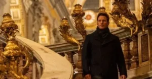 A man in a black coat stands in a grand, ornate setting with golden decorations.