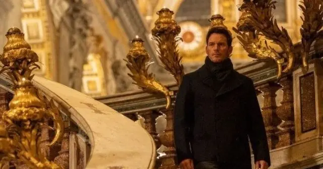 A man in a black coat stands in a grand, ornate setting with golden decorations.