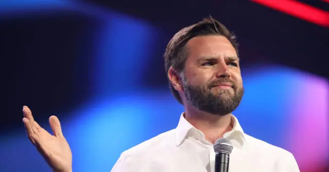 The image shows a man speaking into a microphone, gesturing with his hand, against a colorful, blurred background.