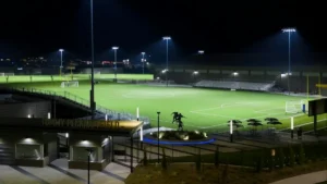 The image shows a well-lit soccer field at night, with a statue and seating area in the foreground, and a cityscape visible in the background.