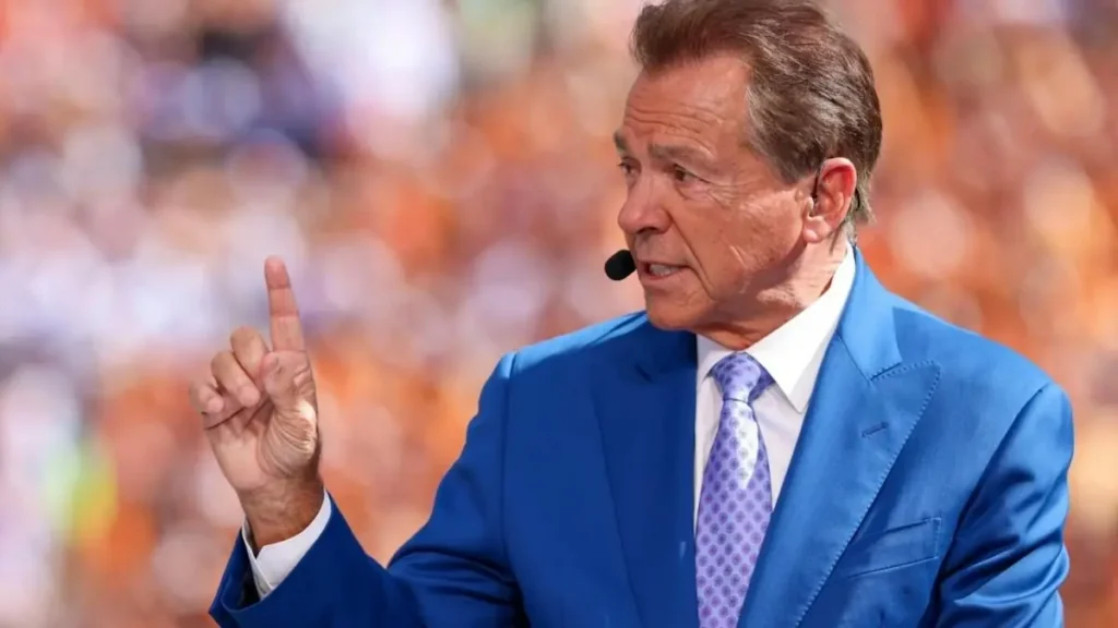 The image shows a man in a blue suit and tie, gesturing with his right hand while speaking into a microphone, likely during a sports broadcast.