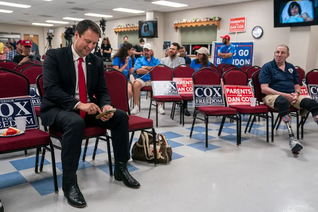 A man in a suit sits on a chair in a campaign setting, surrounded by supporters holding signs for "Cox for Freedom."