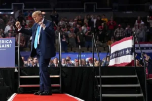 The image shows a political rally with a speaker on stage, accompanied by an American flag and a backdrop that reads "Make America Great Again!"