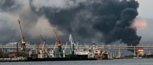 A large plume of black smoke rises from a fire at a port, with cranes and ships in the foreground.