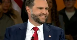 The image shows a man in a suit and red tie, smiling, with an American flag pin on his lapel, in what appears to be a formal setting.