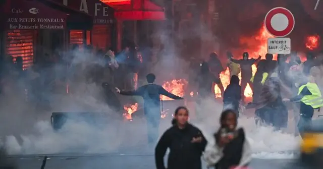 The image depicts a chaotic scene with smoke, fire, and people in a street, suggesting a protest or riot.