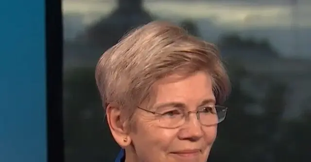 The image shows a person with short, light-colored hair and glasses, wearing a blue top, in what appears to be a professional setting.