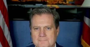 The image shows a man in a suit with an American flag and another flag in the background, suggesting a formal or official setting.