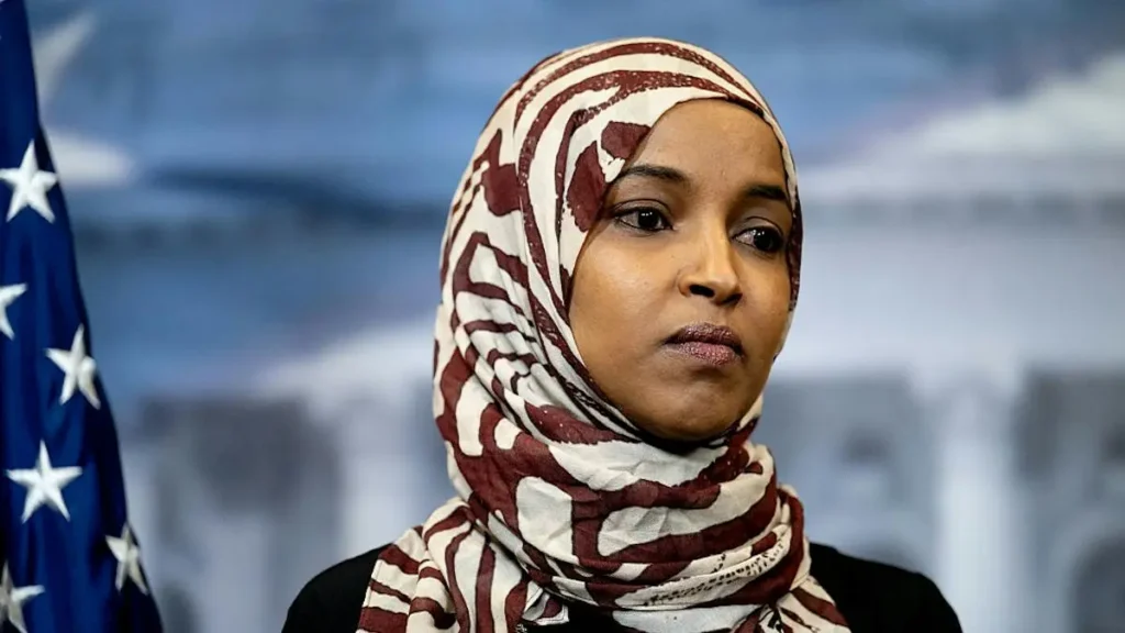 The image shows a person wearing a patterned hijab, with an American flag visible in the background.