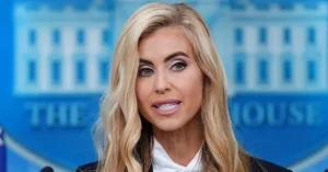 The image shows a person speaking at a press briefing in front of a backdrop featuring the White House logo.