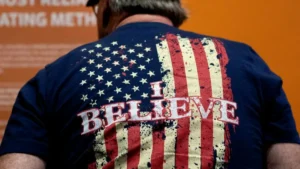 The image shows the back of a person wearing a t-shirt with an American flag design and the text "I BELIEVE" printed on it.