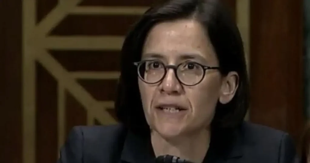 The image shows a person wearing glasses and a suit, speaking in what appears to be a formal setting.