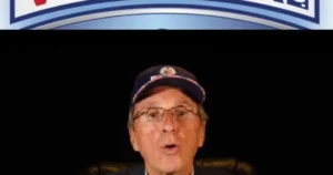 The image shows a man wearing a cap and glasses, speaking in front of a backdrop with a logo.
