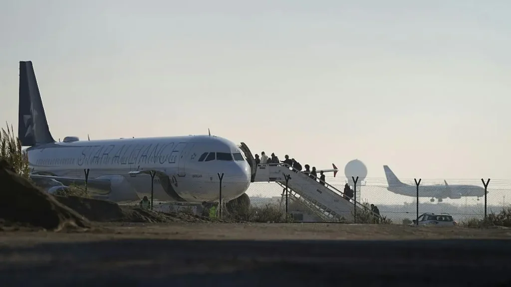 Passengers are boarding a Star Alliance-branded airplane at an airport as another aircraft takes off in the background.
