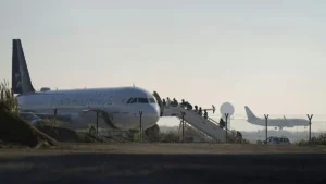Passengers are boarding a Star Alliance-branded airplane at an airport as another aircraft takes off in the background.