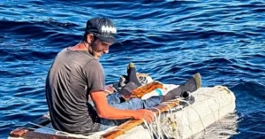 A man sits on a makeshift raft in the middle of the ocean, appearing to be in distress or in need of rescue.