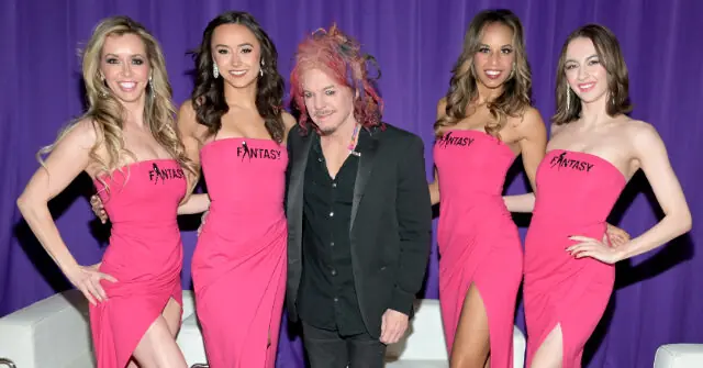 A man stands with four women in matching pink dresses with "Fantasy" written on them, posing for a photo in front of a purple backdrop.