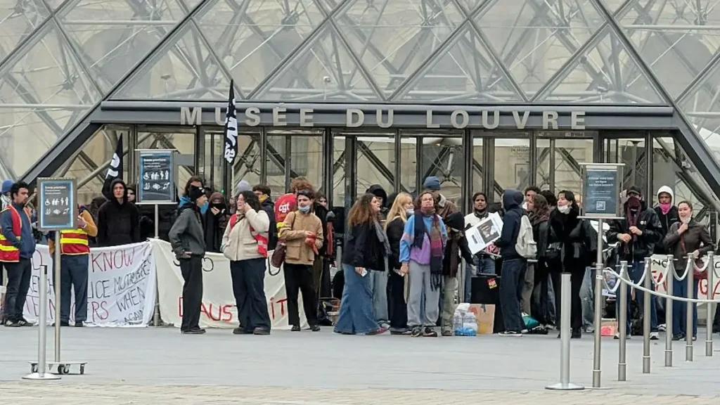 A group of people gather in front of the Louvre Pyramid, holding signs and banners, possibly participating in a protest or demonstration.