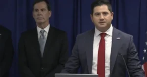 Two men in suits stand at a podium with an American flag in the background, suggesting a formal press conference or official announcement.