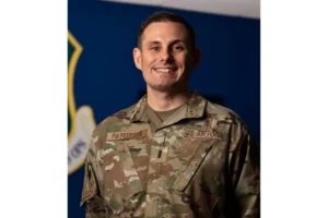The photo shows a U.S. Air Force officer in uniform, standing in front of a blue background with a logo, smiling.