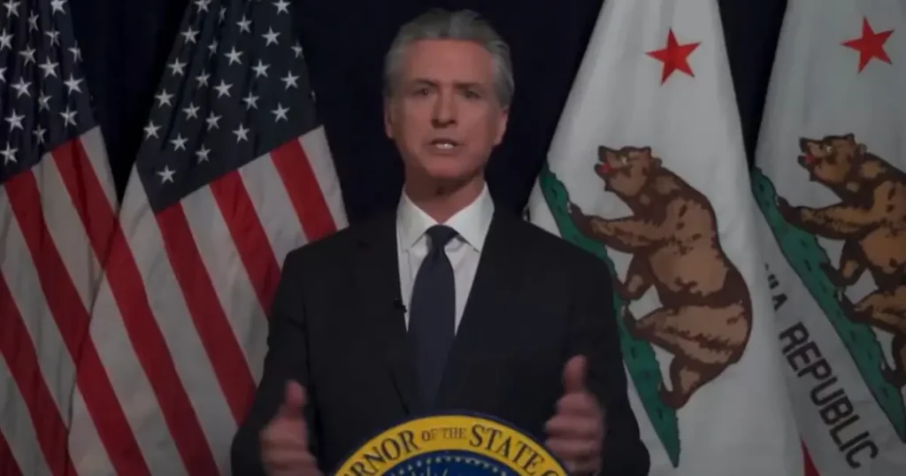 A man in a suit stands at a podium with the seal of the Governor of the State of California and American flags behind him.