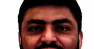 The image shows a close-up of a person with dark hair and a mustache, set against a plain white background.