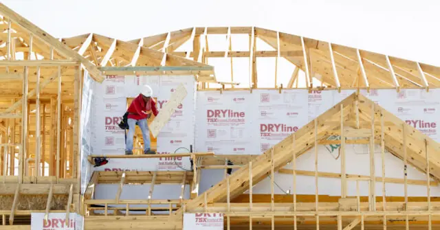 A construction worker is seen working on a house under construction, with wooden beams and insulation visible.