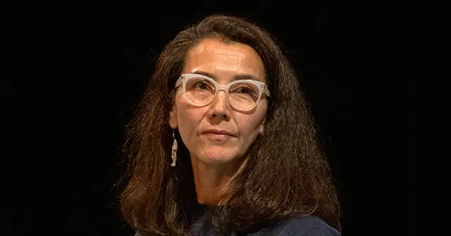The image shows a person with long, dark hair wearing white-framed glasses and a dark top, set against a dark background.