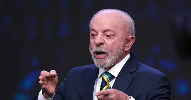 The image shows a man in a suit and tie, gesturing with his hands as he speaks, set against a blurred background.