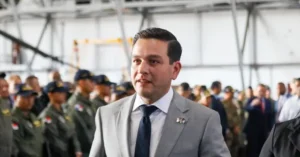 The image shows a man in a suit standing in front of a group of uniformed personnel in a hangar setting.
