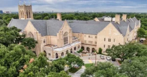 The image shows an aerial view of a large, historic building with a Gothic architectural style, surrounded by trees and a parking area.
