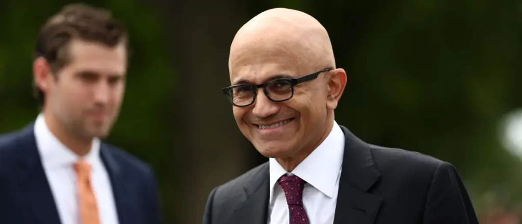 The image shows a man in a suit and tie smiling, with another person partially visible in the background.