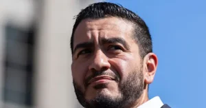 The image shows a close-up of a man with a beard and short hair, wearing a white shirt, against a backdrop of a clear blue sky and a building.
