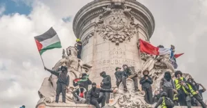 Protesters gather atop a monument, waving flags and holding signs, as part of a demonstration.