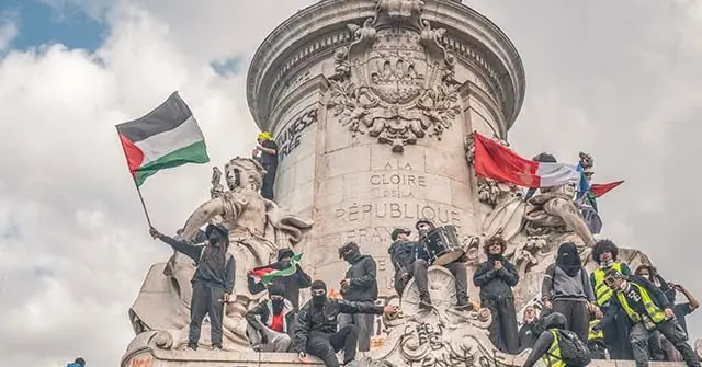 Protesters gather atop a monument, waving flags and holding signs, as part of a demonstration.