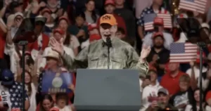 A man in a camouflage shirt and orange hat is giving a speech at a podium in front of a crowd holding American flags and signs.