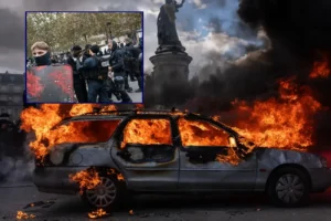 The image depicts a burning car during a protest, with police officers in riot gear and a statue in the background.