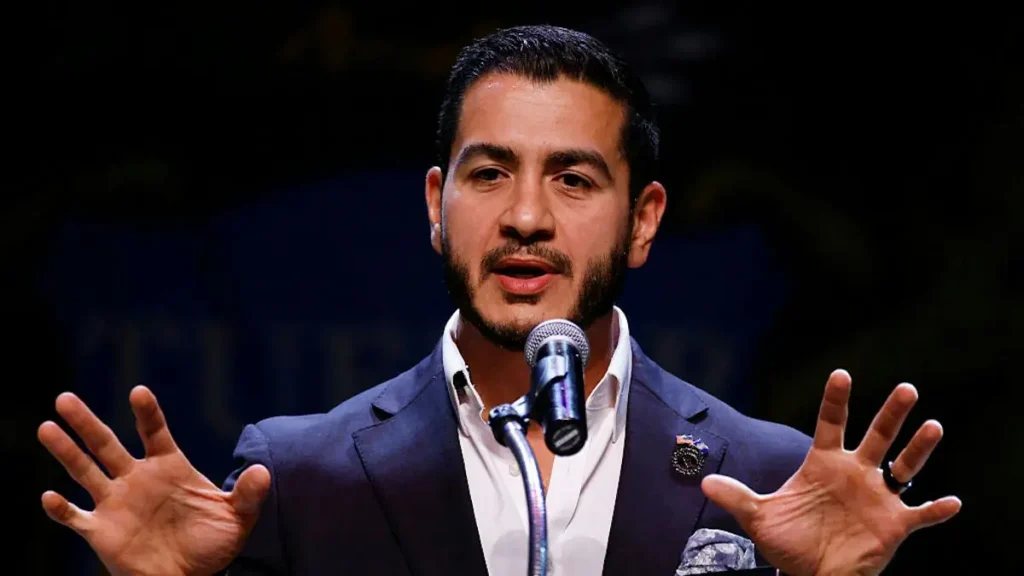 A man in a suit is speaking into a microphone with his hands outstretched, likely addressing an audience.