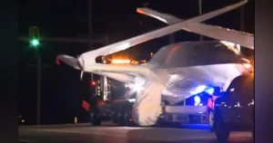 A helicopter is being transported on a flatbed truck at night, with emergency lights visible in the background.