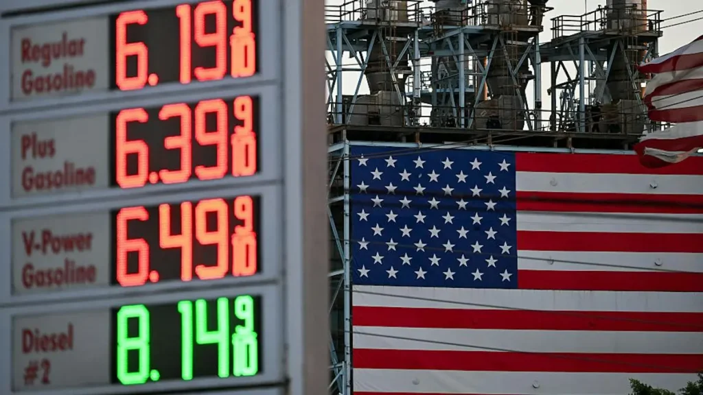 The photo shows a gas price sign displaying high fuel costs alongside an American flag and industrial equipment, symbolizing the economic impact of rising fuel prices.
