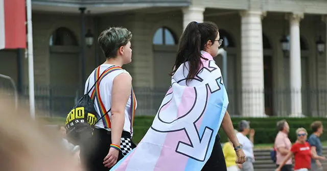 Two individuals walk together, one draped in a transgender flag, symbolizing support and pride for the LGBTQ+ community.