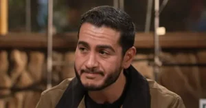The image shows a man with a beard and short hair, wearing a brown jacket and a black shirt, seated in what appears to be a studio setting.