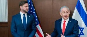 Two individuals in formal attire are standing in front of the American and Israeli flags, engaged in a discussion.