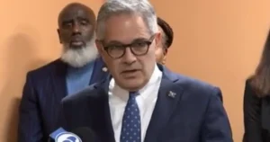 A man in a suit and tie is speaking at a press conference, with other individuals standing behind him.