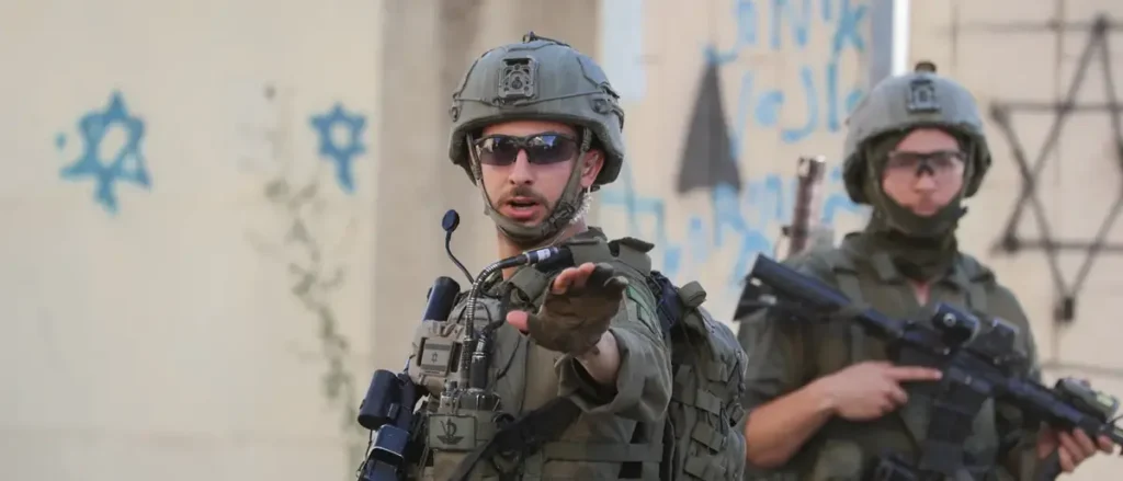 The image shows two soldiers in military gear, one of whom is gesturing with his hand while speaking, with a wall in the background featuring blue Star of David symbols.